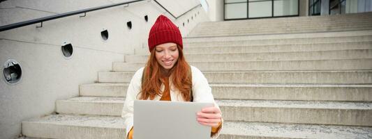 Smiling redhead girl, student sits on stairs outdoors and uses laptop, connects to public wifi in city and works on project, uses internet on computer photo