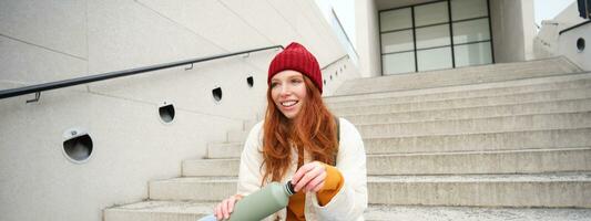 Smiling traveler, redhead girl tourist sits on stairs with flask, drinks hot coffee from thermos while travelling and sightseeing around foreign city, sits on stairs and rests photo