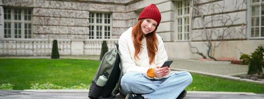 Redhead girl, female student sits with mobile phone on bench in parj, leans on her backpack. Woman browsing social media app feed on her smartphone photo