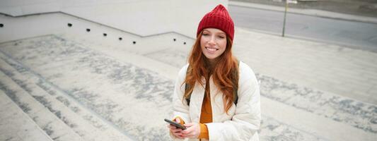 Smiling redhead girl, student tourist walks around city, goes up the stairs, looks at mobile phone map to get around town, sends message on smartphone photo