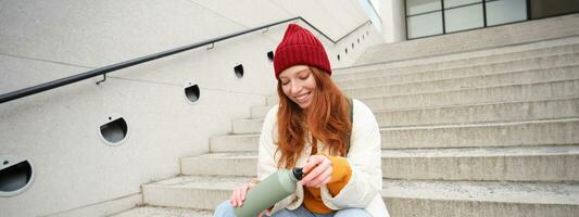 Happy redhead woman, tourist with thermos, drinks her hot tea, coffee from travel flask, restests during her travelling in city and sightseeing photo