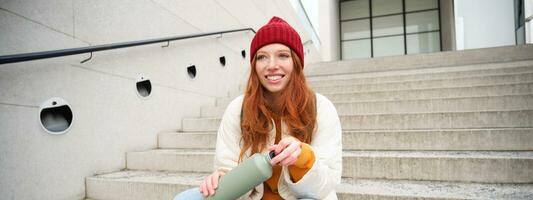 Smiling traveler, redhead girl tourist sits on stairs with flask, drinks hot coffee from thermos while travelling and sightseeing around foreign city, sits on stairs and rests photo