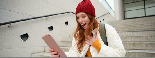 Happy stylish redhead girl, student in red hat, holds digital tablet, uses social media app, searches something online, connects to wifi photo