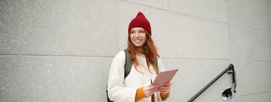 Young redhead girl, walks with digital tablet around city, stands on street with backpack and gadget, looks at online map, searches for a place in internet photo