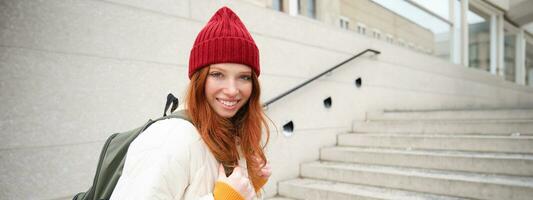 contento joven chica, pelirrojo turista en rojo sombrero con mochila, camina alrededor ciudad, explora ciudad, mochilero alrededor Europa, de viaje solo foto