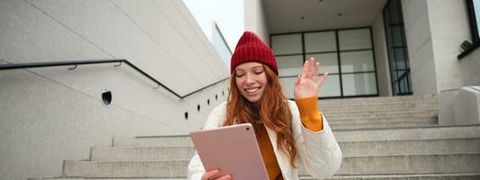 Joyful redhead teen girl, student with digital tablet, says hello, waves hand at gadget camera, connects to video chat, talks to friend in application photo