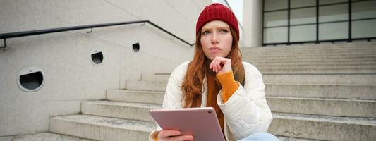 Portrait of troubled redhead girl, college student with thoughtful face, sits on street stairs, holds digital tablet, thinks how to reply on message photo