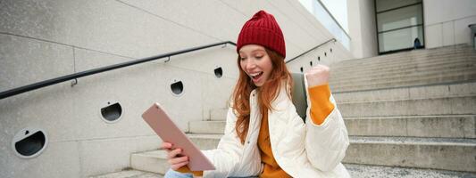 Happy stylish redhead girl, student in red hat, holds digital tablet, uses social media app, searches something online, connects to wifi photo