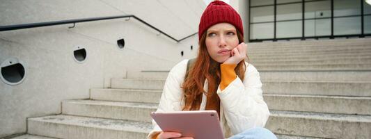 Portrait of troubled redhead girl, college student with thoughtful face, sits on street stairs, holds digital tablet, thinks how to reply on message photo