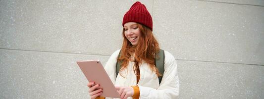 Young redhead girl, walks with digital tablet around city, stands on street with backpack and gadget, looks at online map, searches for a place in internet photo