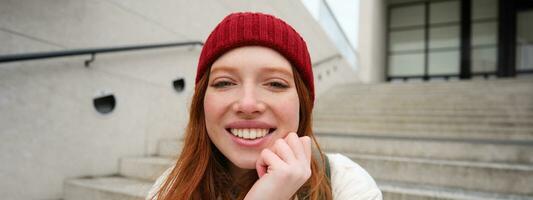 Beautiful redhead student, girl in red hat, smiles sincere, looks happy and relaxed, sits on stairs outdoors photo