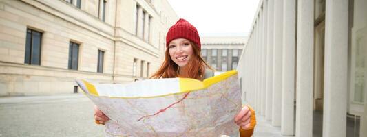 Beautiful redhead woman, tourist with city map, explores sightseeing historical landmark, walking around old town, smiling happily photo