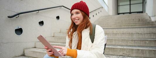 Happy stylish redhead girl, student in red hat, holds digital tablet, uses social media app, searches something online, connects to wifi photo