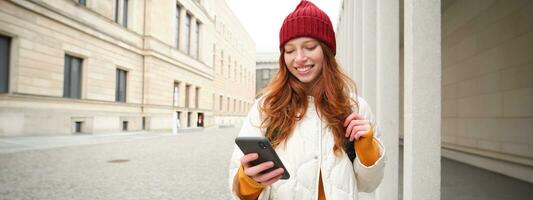 Beautiful smiling girl, tourist with backpack, holding smartphone, using map on mobile phone application, looking for sightseeing in internet app, standing outdoors photo