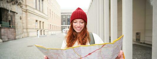 pelirrojo chica, turista explora ciudad, mira a papel mapa a encontrar camino para histórico puntos de referencia, mujer en su viaje alrededor Europa búsquedas para Turismo foto