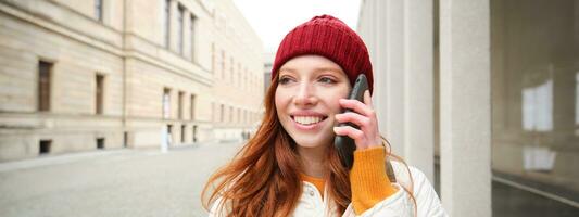 contento pelirrojo mujer, niña con teléfono negociaciones, tiene conversacion en móvil aplicación, usos Internet a llamada extranjero con teléfono inteligente aplicación, riendo y sonriente foto