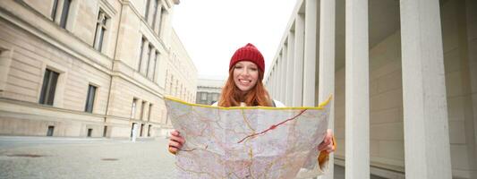 Beautiful redhead woman, tourist with city map, explores sightseeing historical landmark, walking around old town, smiling happily photo
