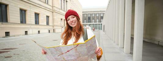 pelirrojo chica, turista explora ciudad, mira a papel mapa a encontrar camino para histórico puntos de referencia, mujer en su viaje alrededor Europa búsquedas para Turismo foto