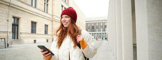 Beautiful smiling girl, tourist with backpack, holding smartphone, using map on mobile phone application, looking for sightseeing in internet app, standing outdoors photo