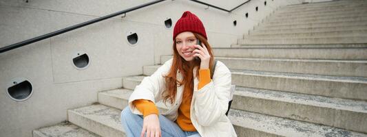 joven elegante pelirrojo niña en rojo sombrero, se sienta en calle y negociaciones en móvil teléfono, tiene teléfono conversación, anillos su amigo mientras relaja al aire libre foto