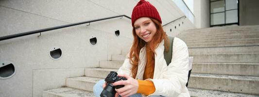 Smiling redhead girl photographer, checks her shots, holds camera and looks at screen, takes photos outdoors, walks around street and does streetstyle shooting