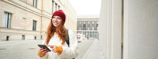 hermosa sonriente chica, turista con mochila, participación teléfono inteligente, utilizando mapa en móvil teléfono solicitud, mirando para Turismo en Internet aplicación, en pie al aire libre foto