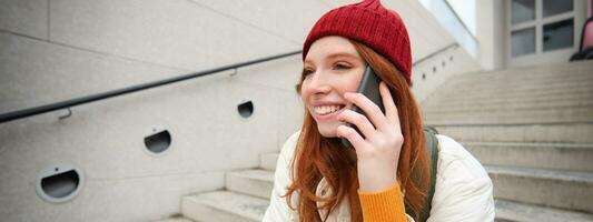 Young stylish redhead girl in red hat, sits on street and talks on mobile phone, has telephone conversation, rings her friend while relaxes outdoors photo