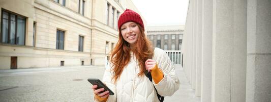 Beautiful smiling girl, tourist with backpack, holding smartphone, using map on mobile phone application, looking for sightseeing in internet app, standing outdoors photo