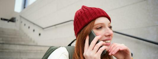 Beautiful smiling redhead female model, sits on street and talks on mobile phone, uses smartphone app to call abroad, laughing during telephone conversation photo