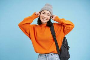 Cute smiling asian girl puts on warm hat to go outside, walks with backpack in orange sweater, blue background photo