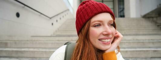Close up portrait of beautiful redhead girl in red hat, urban woman with freckles and ginger hair, sits on stairs on street, smiles and looks gorgeous photo