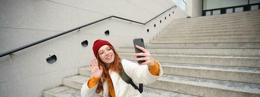 Happy college girl talks with friends on video chat smartphone app, sits on stairs outdoors uses her mobile phone application, waves hand at telephone camera photo