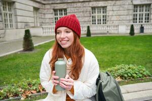 Smiling redhead girl rests in park, sits on bench with backpack, drinks from thermos, enjoys hot drink from flask and looks relaxed photo