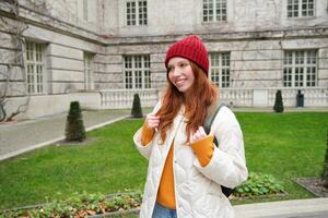 Cute ginger girl in red hat, holds backpack, walks around city and smiles. Student going to campus in University photo