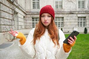 Portrait of confused redhead girl with smartphone, shrugs shoulders with puzzled, clueless face expression, tourist got lost in city photo