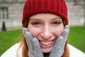 Close up portrait of beautiful redhead woman in red knitted hat, warm gloves, smiling and looking happy at camera, sitting in park photo