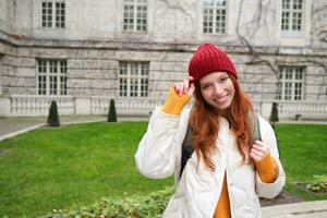 Beautiful redhead woman with backpack, tourist walking around city park and looks around, wears red hat and warm coat photo