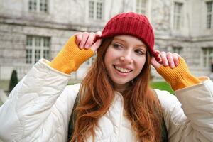 Portrait of smiling redhead woman puts on red hat and smiles, wearing warm clothes while exploring city streets photo