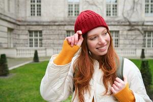 Beautiful redhead woman with backpack, tourist walking around city park and looks around, wears red hat and warm coat photo