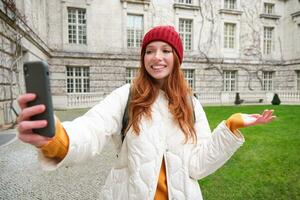 Portrait of happy tourist, girl with backpack, video chats, shows building, sightseeing attraction to friend, poses near landmark and smiles photo