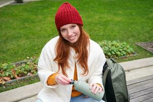 Smiling redhead girl rests in park, sits on bench with backpack, drinks from thermos, enjoys hot drink from flask and looks relaxed photo