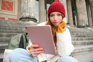 Portrait of young redhead woman sitting outdoors on stairs, reading ebook on digital tablet, wearing red hat and warm clothes photo