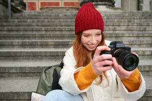 Portrait of young photographer girl, sits on stairs with professional camera, takes photos outdoors, making lifestyle shooting