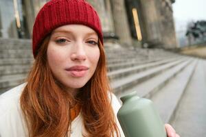 Young redhead female tourist rests during her trip, opens thermos and drinks hot tea, having a break after sightseeing photo