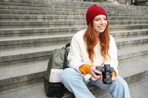 Young student, photographer sits on street stairs and checks her shots on professional camera, taking photos outdoors