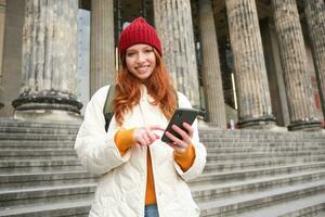 Portrait of young redhead woman holding mobile phone, using smartphone app, walking in city centre, using map application or texting a message photo