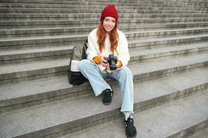 Young student, photographer sits on street stairs and checks her shots on professional camera, taking photos outdoors