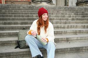 Young redhead female tourist rests during her trip, opens thermos and drinks hot tea, having a break after sightseeing photo