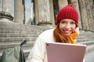 Close up portrait of cute redhead european girl, sitting in red hat near municipal building on stairs, holds digital tablet, reads ebook or surfs internet photo