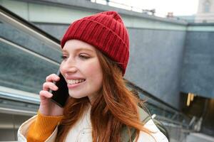 Portrait of happy redhead woman walking around town with smartphone, calling someone, talking on mobile phone outdoors photo